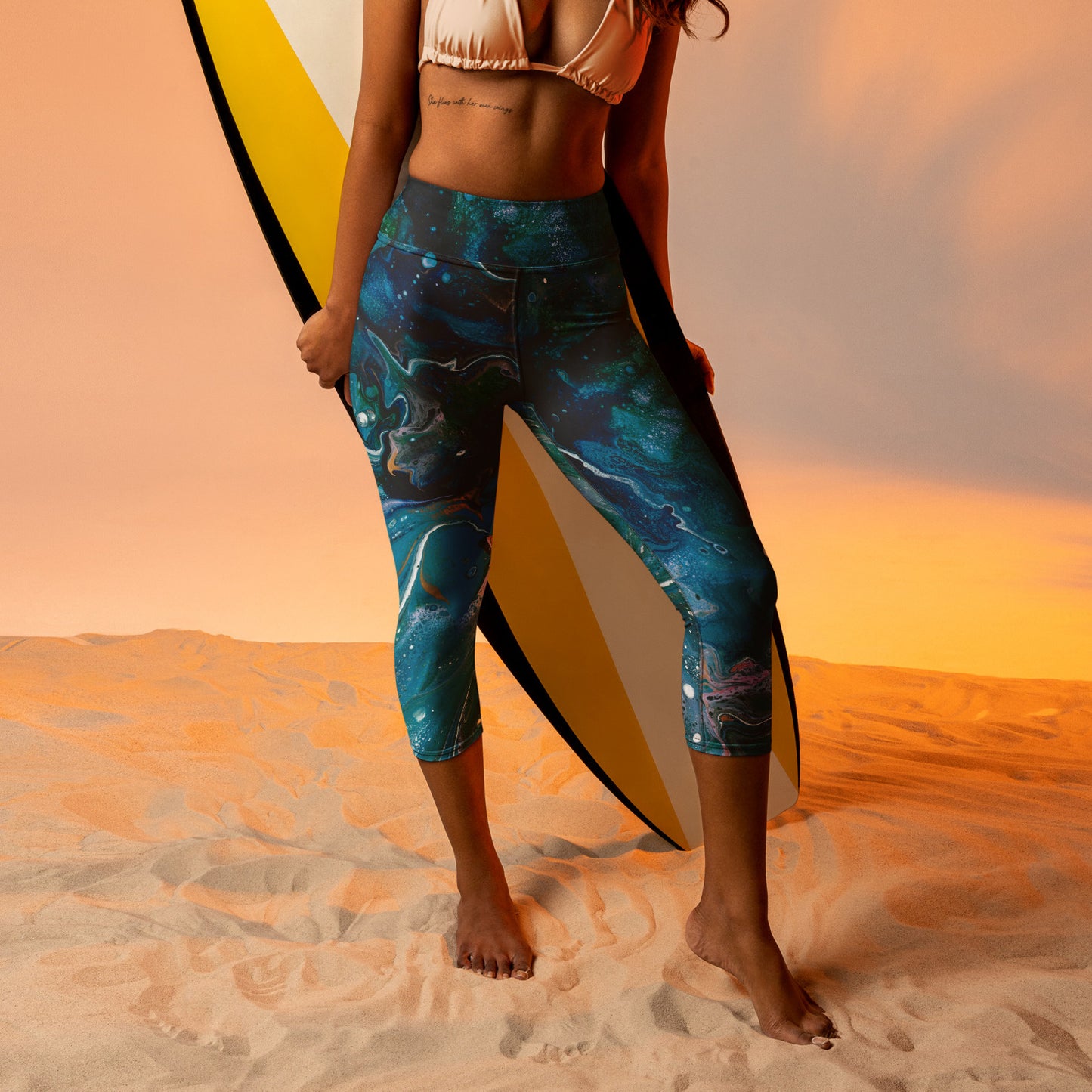 Lifestyle image with model wearing orange string bikini top and these Ultimate Comfort Capri Yoga Pants. Model is holding surfboard on sandy. Front view of pants.